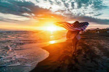 Güzel vatansever, hayat dolu genç bir kadın. Amerikan bayrağı uzanmış ellerinde. Yaz günbatımında ayakta duruyor.