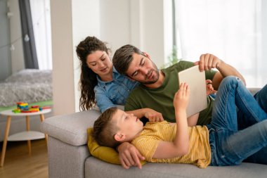 Çocuklu mutlu bir aile tablet bilgisayar kullanarak eğleniyor, ebeveynler ve çocuklar gülüyor dijital cihazla evde dinleniyorlar, komik internet videoları izliyorlar, online arama yapıyorlar.