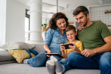 Mutlu aile ve çocuk akıllı telefonla eğleniyor, dijital cihazlar evde. Çocuk telefon oyunu oynamak için uygulama kullanıyor, komik cep telefonu videoları izliyor, kanepede online arama yapıyor.