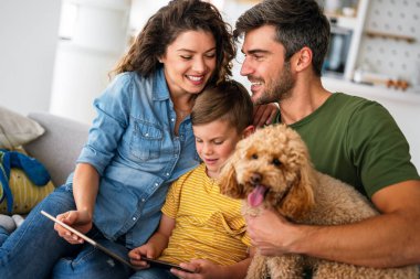 Neşeli Kafkas ailesi evde vakit geçiriyor. Mutlu çocuk tablette video veya çizgi film izlerken ebeveynler evcil köpek yavrusuyla kanepede oynuyorlar.