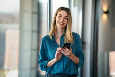 Şirket ofisinde haftalık programı analiz ederken cep telefonu kullanan mutlu bir iş kadını.