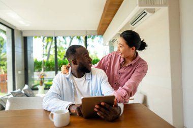 Genç ve çok ırklı çift evde tablet kullanırken eğleniyor. Tablet yazarken kucaklaşan genç insanları sevmek. Mutlu çift gelecek planlarını konuşuyor.