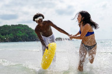 Mutlu ırklar arası bir çift, eğlenceli bir plaj gününün tadını çıkarıyor, okyanus dalgalarında su sıçratıyorlar, el ele tutuşuyorlar, ve sarı yüzme yüzüğüyle güneşli gökyüzünün altında gülümsüyorlar.