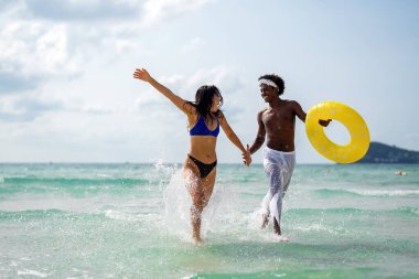 Irklar arası bir çift sahilde eğleniyor, okyanus dalgalarında koşuyor, gülümsüyor ve güneşli bir yaz gününde tropik bir tatilin tadını çıkarıyor.