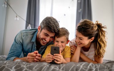 Mutlu aile ve çocuk akıllı telefonla eğleniyor, dijital cihazlar evde. Çocuk telefon oyunu oynamak için uygulama kullanıyor, komik cep telefonu videoları izliyor, kanepede online arama yapıyor.
