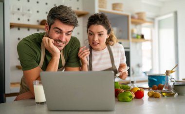 Genç kadın ve erkek online aşçılık atölyesine katılmak için dizüstü bilgisayar kullanıyor, internetten vegan mutfak tarifleri öğreniyorlar. İnternette yemek kursunu izleyen mutlu çift.