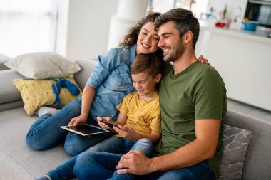 Mutlu aile ve çocuk akıllı telefonla eğleniyor, dijital cihazlar evde. Çocuk telefon oyunu oynamak için uygulama kullanıyor, komik cep telefonu videoları izliyor, kanepede online arama yapıyor.