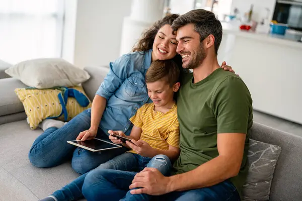 Mutlu aile ve çocuk akıllı telefonla eğleniyor, dijital cihazlar evde. Çocuk telefon oyunu oynamak için uygulama kullanıyor, komik cep telefonu videoları izliyor, kanepede online arama yapıyor.