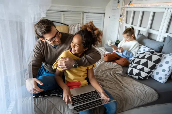 Ergen kızıyla birlikte modern tablet kullanan, evdeki rahat yatakta oturan baba ve sevimli Afro-Amerikan kız çocuğu cihaz ekranına bakıyor, oyun oynuyorlar.
