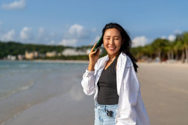 Genç Asyalı mutlu bir kadın tropik bir plajda akıllı telefonuyla konuşuyor tatilden, uzaktan işten, sosyal medyadan ve kaygısız bir dijital yaşam tarzından zevk alıyor.