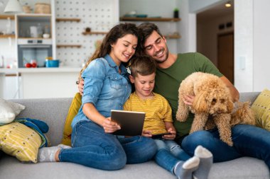 Neşeli Kafkas ailesi evde vakit geçiriyor. Mutlu çocuk tablette video veya çizgi film izlerken ebeveynler evcil köpek yavrusuyla kanepede oynuyorlar.
