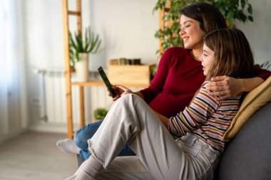 Çekici gülümseyen anne genç kızıyla kanepede rahatlar, cep telefonunu tutar, sosyal medya ağında sohbet eder, komik videolar izler, yeni modern uygulamalar öğrenir, hafta sonu evde internetin tadını çıkarır.