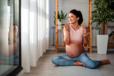 Mutlu hamile bir kadın akıllı telefonunu kullanıyor ve evde egzersiz yaparken selfie çekiyor..