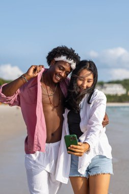 Tropikal bir plajda selfie çeken, tatilin tadını çıkaran, sosyal medya içeriği yaratan ve yaz güneşi altında dijital göçebe yaşam tarzı olan mutlu çiftler.