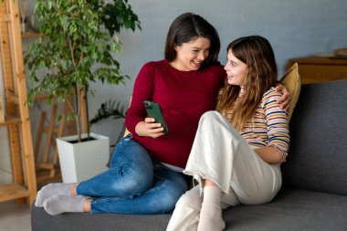 Çekici gülümseyen anne genç kızıyla kanepede rahatlar, cep telefonunu tutar, sosyal medya ağında sohbet eder, komik videolar izler, yeni modern uygulamalar öğrenir, hafta sonu evde internetin tadını çıkarır.