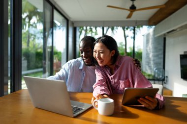 Çok uluslu çift sabah rutininin keyfini çıkarıyorlar, gülümsüyorlar ve rahat ve modern bir ev ortamında dizüstü bilgisayar ve tablet kullanıyorlar.