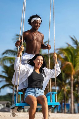 Mutlu ırklar arası bir çift, eğlenceli bir plaj gününün tadını çıkarıyor, palmiye ağaçlarının altında sallanıyor, gülüyor ve kaygısız hissediyorlar güneş ışığı ve mavi gökyüzü ile tropik bir tatilde.