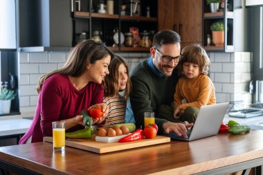Evde dizüstü bilgisayar kullanan heyecanlı çocukları olan mutlu genç ebeveynler. Video konferansında sanal parti vermek, ekommerce ailesinin online alışverişini yapmak..
