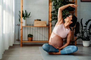 Aktif hamile kadın yoga yapıyor, kaslarını mindere geriyor, hamilelik öncesi egzersizleri yapıyor ve vücudu doğuma hazırlıyor.