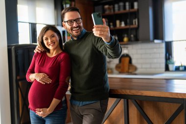 Sevimli bir adam hamile karısına sarılıyor, hamileliğinin son üç ayında selfie çekerek eğleniyor. Mutlu koca hamile kadını kucaklarken bebek bekliyor..