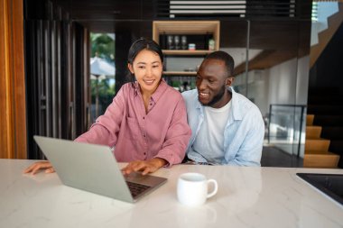 Çevrimiçi alışveriş veya ödeme için dijital dizüstü bilgisayar kullanan mutlu çift. Kadın kocasına dijital cihazdan bir şey alıyor. Orta Doğulu, Afro-Amerikalı çift dinleniyor..