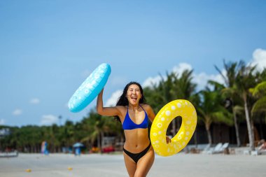 Heyecanlı genç kadın tropikal plaj tatilinin tadını çıkarıyor, elinde renkli şişirilebilir yüzükler tutuyor ve eğlenceli bir yaz kaçamağını kutluyor.