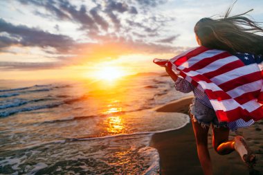 Gülümseyen ve sahilde koşan mutlu kadın. Amerikan bayrağı taşıyan bir kadının portresi. 4 Temmuz bağımsızlık günü. ABD 'de tatil. ABD sembolü. Gün batımı
