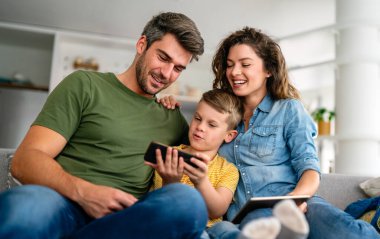 Mutlu aile ve çocuk akıllı telefonla eğleniyor, dijital cihazlar evde. Çocuk telefon oyunu oynamak için uygulama kullanıyor, komik cep telefonu videoları izliyor, kanepede online arama yapıyor.