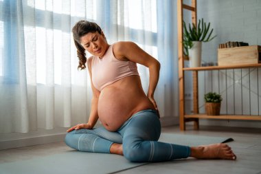 Hamile kalırken bel ağrısından muzdarip genç bir anne. Hamile bir kadın, sırtında ağrı hissederek ve rahatlatıcı fizyoterapi egzersizleri yaparak yorgun düşüyor.