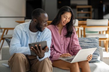 Çok uluslu çift sabah rutininin keyfini çıkarıyorlar, gülümsüyorlar ve rahat ve modern bir ev ortamında dizüstü bilgisayar ve tablet kullanıyorlar.