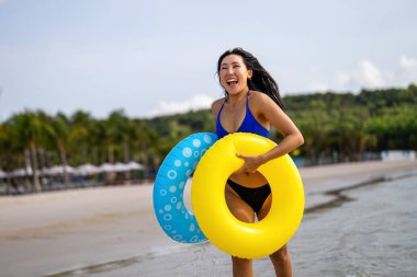 Heyecanlı genç kadın tropikal plaj tatilinin tadını çıkarıyor, elinde renkli şişirilebilir yüzükler tutuyor ve eğlenceli bir yaz kaçamağını kutluyor.