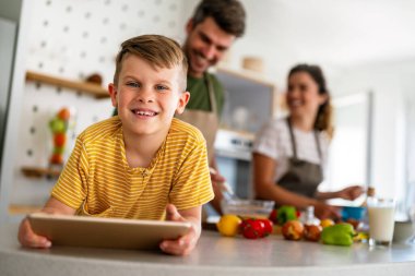 Mutfakta çocuklu mutlu bir aile birlikte modern tablet kullanarak eğleniyor, gülümseyen ebeveynler çocuklarla birlikte hafta sonunun keyfini çıkarıyor evde bilgisayardan gülen videolar izliyorlar.