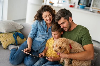 Çocuklu mutlu bir aile tablet bilgisayar kullanarak eğleniyor, ebeveynler ve çocuklar gülüyor dijital cihazla evde dinleniyorlar, komik internet videoları izliyorlar, online arama yapıyorlar.