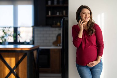 Mutlu, rahat, hamile bir kadın cep telefonu kullanıyor, elinde akıllı telefonuyla gülen bir kadın, cep telefonuna bakıyor, mobil uygulamalarda online ekommerce alışverişi yapıyor ya da video seyrediyor.