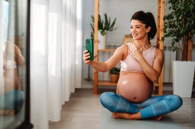 Mutlu hamile bir kadın akıllı telefonunu kullanıyor ve evde egzersiz yaparken selfie çekiyor..