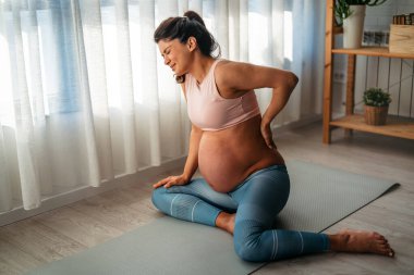 Braxton Hicks kasılmaları. Genç hamile bir kadın yoga egzersizi sırasında acı çekiyor. Evde karın ağrısı çekiyor. Mindere oturmuş, karnına dokunuyor ve kramp giriyor..