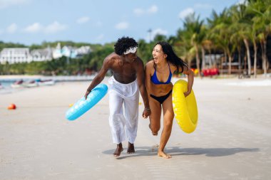 Irklar arası bir çift sahilde eğleniyor, okyanus dalgalarında koşuyor, gülümsüyor ve güneşli bir yaz gününde tropik bir tatilin tadını çıkarıyor.