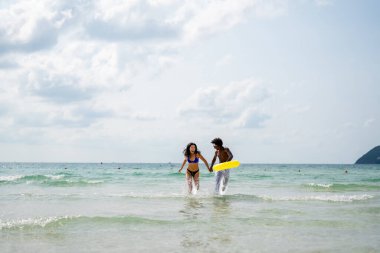 Irklar arası bir çift sahilde eğleniyor, okyanus dalgalarında koşuyor, gülümsüyor ve güneşli bir yaz gününde tropik bir tatilin tadını çıkarıyor.