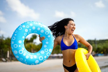 Özgür hissediyorum. Neşeli genç Asyalı bir kadın renkli şişme yüzüklerle ellerini kaldırarak mükemmel bir plaj tatilinin özgürlüğünü kutluyor.