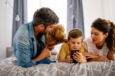 Mutlu aile ve çocuk akıllı telefonla eğleniyor, dijital cihazlar evde. Çocuk telefon oyunu oynamak için uygulama kullanıyor, komik cep telefonu videoları izliyor, kanepede online arama yapıyor.
