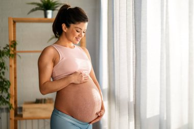Göbeğini gösteren rahat giysiler içinde güzel bir gülümseme ve çatlaklarla dolu mutlu ve formda hamile bir kadın.