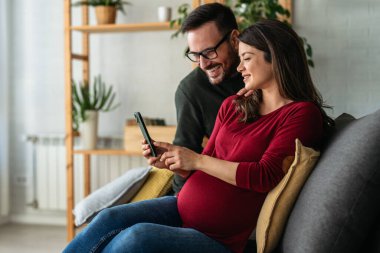 Mutlu genç çift sanal mobil servis kullanıyor, online uygulama akıllı telefonda, oturuyor, koltukta sarılıyor, video görüşmesi yapıyor, internetten alışveriş yapıyor. Hamile kadın ve koca büyük göbek tutuyorlar.