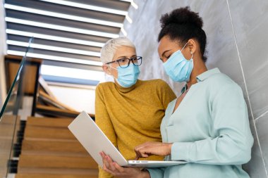 Modern ofiste maske takan iş kadınları, laptopla takım çalışması, salgın sırasında profesyonel iletişim ve işyeri güvenliği