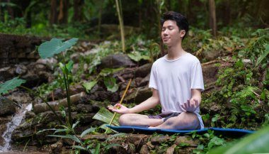 İnsan meditasyon yapıyor, yağmur ormanlarındaki şelalenin yakınındaki kayalıklarda oturuyor. Spor, yoga, pilates, fitness, sağlıklı yaşam tarzı konsepti.