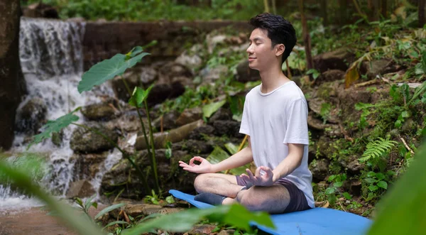Calm man meditating and sitting in lotus pose on the rocks near waterfall in forest. Concept of calm and meditation.