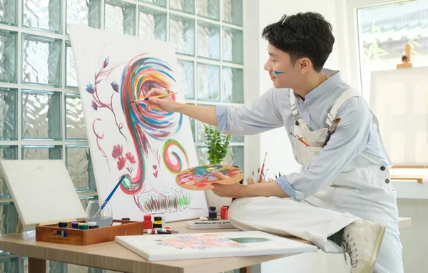 stock image Young man holding paint brushes and palette, painting on canvas at studio.