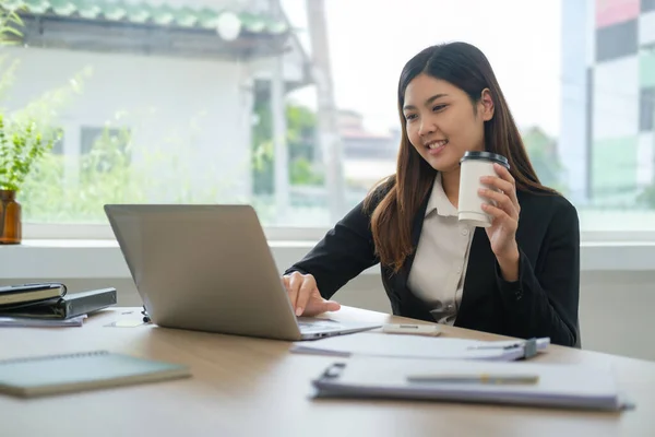 女性従業員のコーヒーを飲むと明るいオフィスでノートパソコンを使用して笑顔 — ストック写真