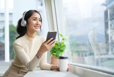 Güzel bir kadın rahat bir odada pencerenin yanında dururken kulaklık takıyor..