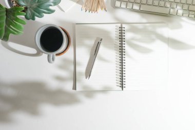 Boş not defteri, kalem, kahve fincanı, ev bitkisi ve klavyenin beyaz masadaki üst görünümü.