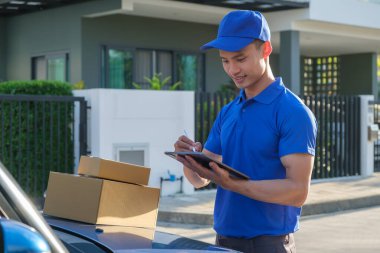 Mavi üniformalı Asyalı erkek kurye dijital tablet üzerine bir paket teslim etmek için bilgileri kontrol ediyor.